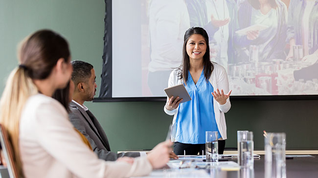 A woman stands at the head of a conference table making a presentation to people seated at the table.