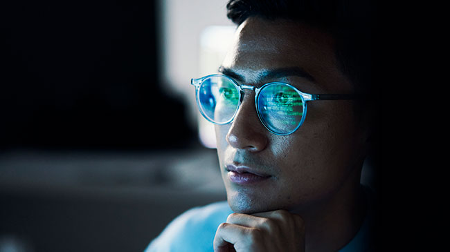 A man is pictured with a computer screen reflected in his eyeglasses.