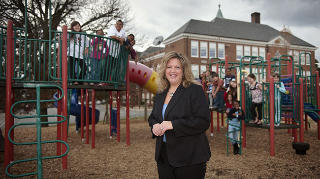 A woman standing in a playground with a lot of children playing in the background