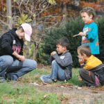 Three kids look towards an adult as they conduct a science experiment outdoors