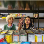 Two women stand among shelves of canned food.