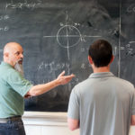 A professor explaining a mathematics problem to a student while standing in front of a blackboard