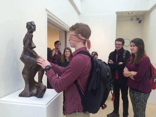 A blindfolded student touches a sculpture while his classmates look on.