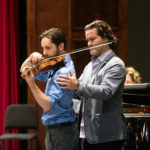 A teacher has his hand on a student's shoulder as he plays the violin