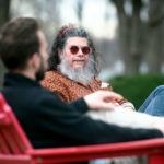 A middle-aged man with sunglasses and a white beard sitting outdoors in a red Adirondack chair talking with a younger, dark-haired man who has his back to the camera