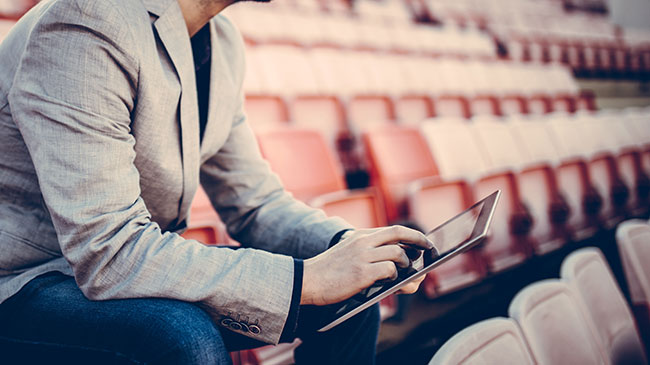 A man sits in the empty bleachers of a sports stadium, holding a tablet