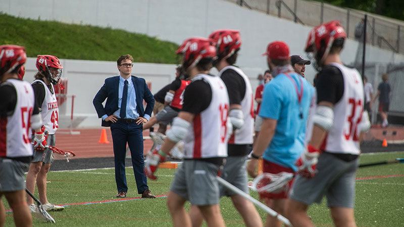 A man in a suit stands on the sidelines watching a lacrosse game.